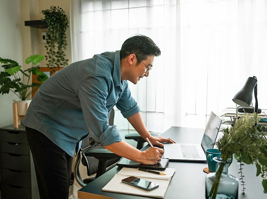 A man writing on paper while looking at two laptops.