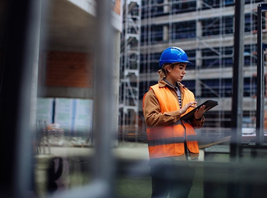 A man in an orange vest and blue helmet holding a tablet.